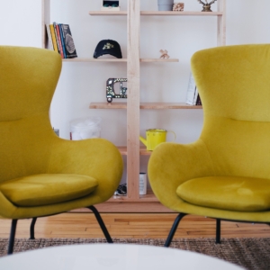 Zwei moderne gelbe Sessel stehen vor einem weißen runden Tisch. Dahinter steht ein Holzregal mit Büchern, einer Pflanze, einer Uhr, einer gelben Teekanne und verschiedenen Dekorationsartikeln. Das Ambiente wirkt gemütlich und modern.