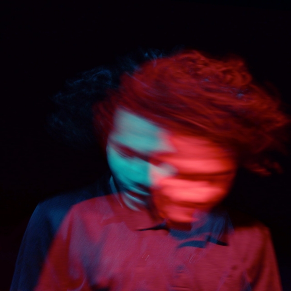 Ein unscharfes Bild einer Person mit lockigem Haar, das durch Bewegung und farbige Lichter in Blau und Rot entsteht und einen geisterhaften Doppelbelichtungseffekt vor einem dunklen Hintergrund erzeugt.