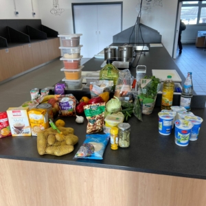 Verschiedene Lebensmittel wie Gemüse, Milchprodukte, Speiseöl, abgepackte Lebensmittel und Gewürze sind auf einer Küchentheke in einer geräumigen, modernen Küche mit Kochstationen und Geräten im Hintergrund angeordnet.