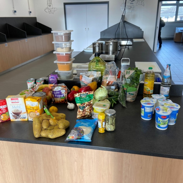 Verschiedene Lebensmittel wie Gemüse, Milchprodukte, Speiseöl, abgepackte Lebensmittel und Gewürze sind auf einer Küchentheke in einer geräumigen, modernen Küche mit Kochstationen und Geräten im Hintergrund angeordnet.