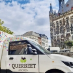 Ein weißer Lieferwagen mit bunten Tupfen und dem Logo QUERBEET AACHEN parkt in der Nähe historischer Gebäude mit Türmen und kunstvoller Architektur in Aachen, Deutschland, unter einem teilweise bewölkten Himmel.