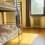 Ein sonnendurchfluteter Raum mit Holzfußboden, einem Etagenbett aus Metall mit weißem Bettzeug, einem Schrank, einem Fenster mit braunen Vorhängen und einem Heizkörper unter dem Fenster.