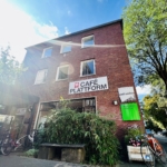 Ein dreistöckiges Backsteingebäude beherbergt das Café Plattform, über dem Eingang prangt ein weißes Schild. Draußen sind Fahrräder geparkt, der Eingang ist begrünt, und das Sonnenlicht fällt durch einen Baum unter einem strahlend blauen Himmel.