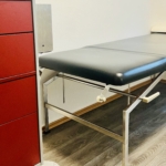 Ein medizinischer Untersuchungstisch mit einem schwarzen Kissen und einer Papierrolle an der Kopfstütze steht neben einem hohen roten Schrank in einem sauberen, hellen Raum mit Holzfußboden.