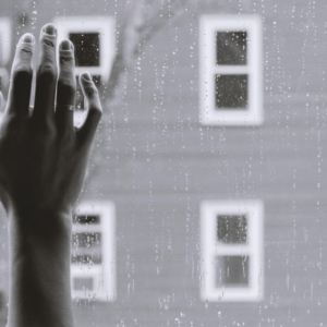 Eine Hand mit gespreizten Fingern berührt ein regengesprenkeltes Fenster, während draußen im Hintergrund unscharfe Gebäude und Fenster zu sehen sind, was eine Stimmung der Sehnsucht oder Kontemplation erzeugt.