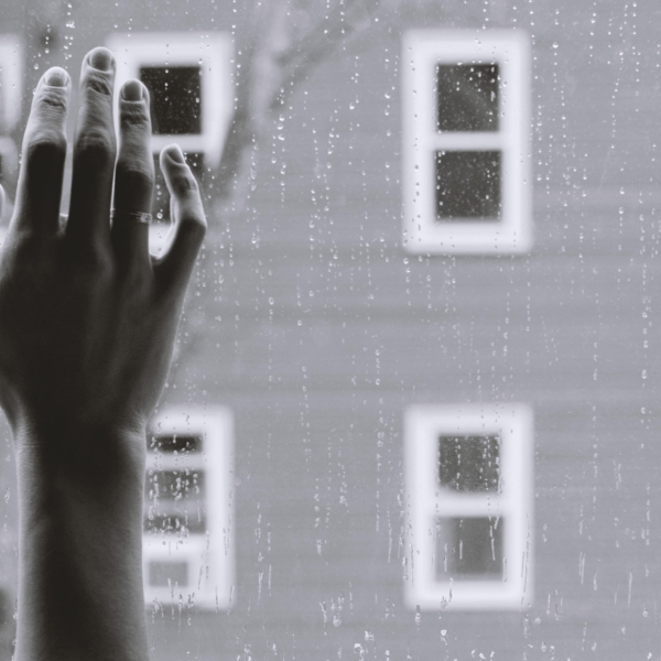 Eine Hand mit gespreizten Fingern berührt ein regengesprenkeltes Fenster, während draußen im Hintergrund unscharfe Gebäude und Fenster zu sehen sind, was eine Stimmung der Sehnsucht oder Kontemplation erzeugt.