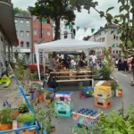 Eine lebhafte Straßenszene mit Menschen, die sich unter einem weißen Vordach versammelt haben, umgeben von bunten Pflanzgefäßen aus bemalten Dosen und Kisten. Pflanzen und Blumen säumen den Bürgersteig, und im Hintergrund stehen Gebäude und Bäume.