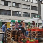 Menschen versammeln sich auf einem Straßenmarkt im Freien mit Holzpaletten als Tische, bunten Schildern und Blumenarrangements. Eine Frau steht in der Mitte, und eine Gruppe sitzt unter einem Zelt. Im Hintergrund ist ein graues Gebäude zu sehen.