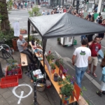 Eine Straßenszene zeigt einen Marktstand unter einem Vordach mit Pflanzen und bunten Schildern auf einem Holztisch. Menschen stehen und unterhalten sich in der Nähe; ein Mann sitzt lächelnd da. Im Hintergrund wartet eine Menschenmenge auf der anderen Straßenseite.