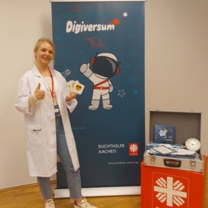 Eine lächelnde Frau in einem weißen Laborkittel steht neben einem Banner mit einem Cartoon-Astronauten und dem Wort "Digiversum". Sie hält Aufkleber in der Hand und zeigt den Daumen nach oben. Ein Koffer und eine Uhr mit einem Caritas-Logo stehen in der Nähe auf dem Boden.