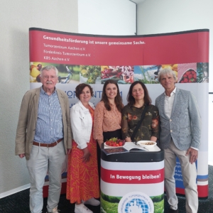 Fünf Personen, drei Frauen und zwei Männer, stehen lächelnd vor einem Display zur Gesundheitsförderung mit Bannern und einem kleinen Tisch mit Obst. Im Hintergrund ist deutschsprachiges Werbematerial zu sehen.