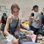 Eine Gruppe von Menschen versammelt sich um eine bunte, mit Pflanzen und Tischen geschmückte Treppe und besucht eine Veranstaltung oder einen Markt im Freien. Über der Treppe hängen bunte Fahnen, und einige Leute beobachten, stöbern oder unterhalten sich in der lebhaften Atmosphäre.