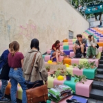 Die Menschen versammeln sich auf den farbenfrohen, pastellfarbenen, mit Pflanzen und Gartenartikeln geschmückten Blockstufen. Einige sitzen und unterhalten sich, während andere in der Nähe stehen. In der Mitte ist ein handgefertigtes "PflanzenTausch"-Schild zu sehen.