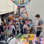 Eine Gruppe von Menschen versammelt sich auf bunten, gekachelten Stufen in einem städtischen Außenbereich und nimmt an einem Pflanzenworkshop teil. Tische mit Pflanzen und Werkzeugen sind aufgestellt, und über den Köpfen hängen leuchtende Transparente.