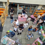 Eine Gruppe von Menschen versammelt sich auf einem Markt oder einer Messe im Freien, von oben gesehen. Bunte Tische, Blumen und Kunstgegenstände liegen verstreut herum. Einige Leute unterhalten sich, andere betrachten die Artikel auf den Tischen, und ein großes OPEN-Banner ist zu sehen.