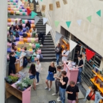 Eine lebhafte Veranstaltung unter freiem Himmel auf einem abgesenkten Platz mit bunten Stufen, die mit Menschen gefüllt sind. Stände mit Blumen, rosa Tische und festliche Wimpel schmücken den Platz. Über der Treppe hängt ein Transparent mit der Aufschrift "ÜBERBEET".