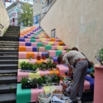 Eine Person arrangiert Topfpflanzen auf bunten, bemalten Stufen neben einer schlichten Treppe. Hilfsmittel und Werkzeuge sind in der Nähe verstreut. Die Stufen sind mit einem Regenbogenmuster versehen und darüber hängen festliche Fahnen.
