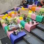Bunte Stufen mit Topfpflanzen, Gartengeräten, Erdsäcken und einem Schild mit der Aufschrift PFLANZENTAUSCH. Oben auf der Treppe sitzen mehrere Menschen, die sich an einer mit Wandmalerei beschichteten Wand unterhalten.