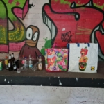 Zwei bunte Einkaufstüten und verschiedene leere Flaschen und Dosen stehen auf einem Betonsims vor einer mit bunten Graffiti überzogenen Wand.