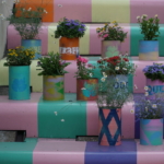 Bunte Topfblumen, die auf einer pastellfarbenen Treppe angeordnet sind, wobei jede Stufe einen anderen zarten Farbton aufweist. Die Töpfe sind mit verschiedenen Mustern und Farben verziert und verleihen der Szene einen lebendigen, künstlerischen Touch.