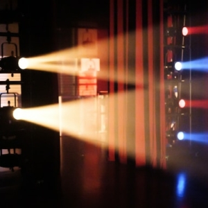 Bühnenscheinwerfer werfen rote, blaue und weiße Lichtstrahlen durch Dunst auf eine abgedunkelte Theaterbühne, während im Hintergrund Geräte und Lichtstrukturen zu sehen sind.