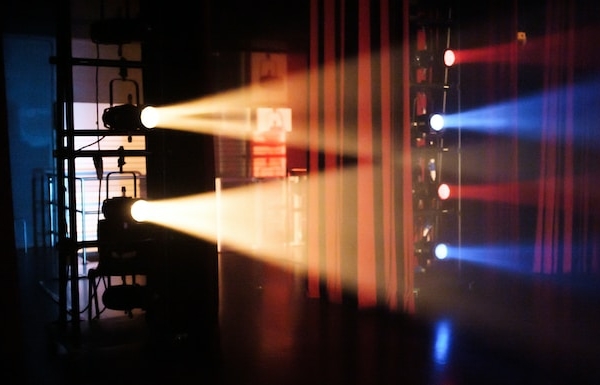Bühnenscheinwerfer werfen rote, blaue und weiße Lichtstrahlen durch Dunst auf eine abgedunkelte Theaterbühne, während im Hintergrund Geräte und Lichtstrukturen zu sehen sind.