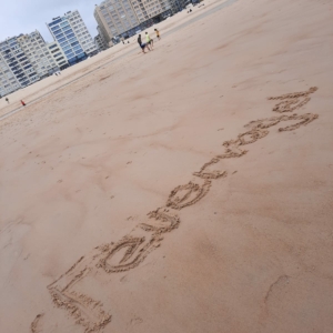 Das Wort Te extraño! ist in den Sand eines weiten, leeren Strandes geschrieben. In der Ferne sind ein paar Menschen zu sehen, die spazieren gehen, und im Hintergrund stehen hohe Wohnhäuser unter einem bewölkten Himmel.