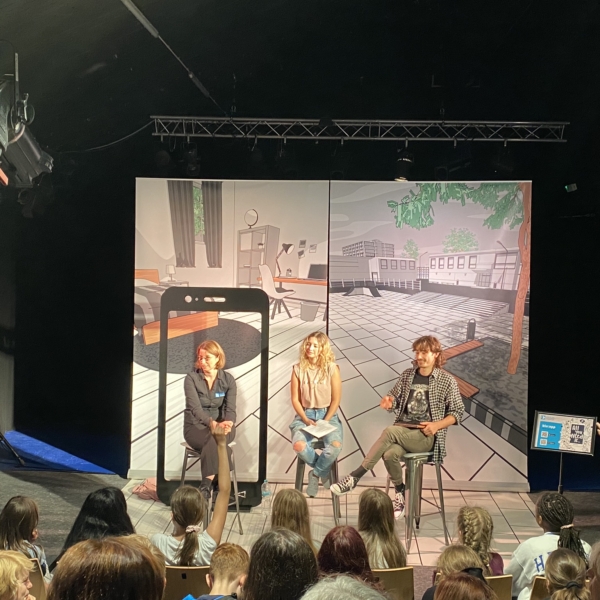 Drei Personen sitzen auf Hockern auf der Bühne vor einer Kulisse, die eine Schulszene darstellt, und sprechen zu einem Publikum. Das Publikum besteht hauptsächlich aus jungen Leuten. Über ihnen und um sie herum sind Bühnenbeleuchtung und Tontechnik zu sehen.
