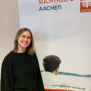 Eine lächelnde Frau mit Brille und schwarzem Pullover steht neben einem Transparent der Suchthilfe Aachen. Das Banner zeigt eine Person von hinten und enthält das Caritas-Logo und die Website-URL.