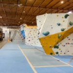 Eine Indoor-Kletterhalle mit großen Boulderwänden mit bunten Griffen. Die Wände haben verschiedene Winkel und Routen, der Boden ist zur Sicherheit blau gepolstert. Eine Person sitzt am anderen Ende der Halle.