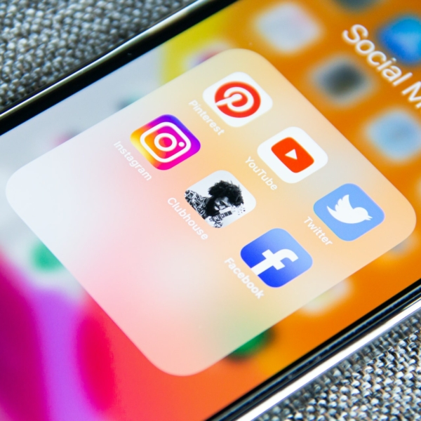 Ein Smartphone-Bildschirm zeigt einen Ordner mit der Bezeichnung "Social Media" mit sechs App-Symbolen: Pinterest, Instagram, Clubhouse, YouTube, Twitter und Facebook auf einem verschwommenen bunten Hintergrund.