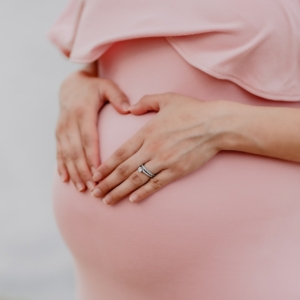 Eine schwangere Frau in einem rosafarbenen Kleid formt mit ihren Händen sanft eine Herzform über ihrem Babybauch. Nur ihr Oberkörper und ihre Hände sind sichtbar.
