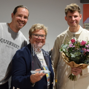 Drei Personen lächeln in die Kamera; die Frau in der Mitte hält eine Trophäe, der Mann auf der linken Seite hebt feierlich die Faust, und der Mann auf der rechten Seite hält einen Blumenstrauß. Im Hintergrund ist ein buntes Plakat zu sehen.