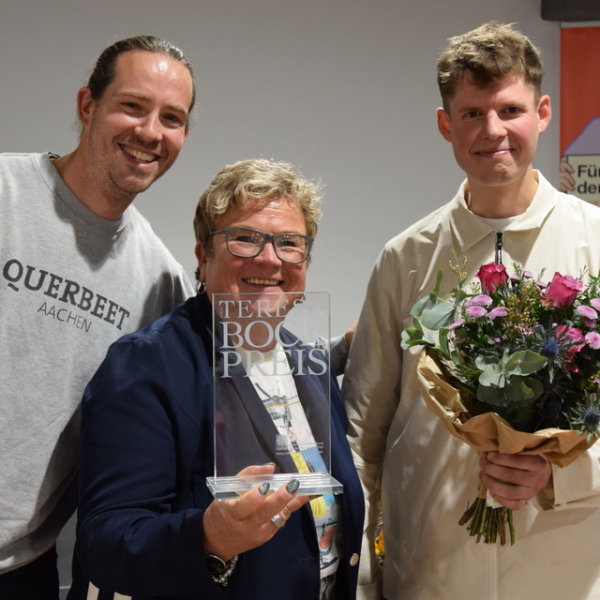Drei Personen lächeln in die Kamera; die Frau in der Mitte hält eine Trophäe, der Mann auf der linken Seite hebt feierlich die Faust, und der Mann auf der rechten Seite hält einen Blumenstrauß. Im Hintergrund ist ein buntes Plakat zu sehen.