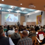 Ein Redner hält einen Vortrag vor einem Publikum, das in einem modernen Konferenzraum sitzt, auf dessen großer Projektionsfläche eine lächelnde Frau zu sehen ist. Die Teilnehmer halten Papiere in der Hand, schauen nach vorne und hören aufmerksam zu.