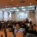 Eine Gruppe von Personen sitzt einem Redner gegenüber, der in einem modernen, gut beleuchteten Konferenzraum einen Vortrag hält, während im vorderen Teil des Raums ein großer Bildschirm ein Foto und einen Text anzeigt.