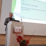 Ein Mann steht an einem Podium und hält einen Vortrag in deutscher Sprache. Auf der projizierten Folie steht: Du bist mir wichtig! Motivierende Gesprächsführung mit konsumierenden Jugendlichen. Vor dem Podium steht ein Blumengesteck.