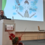 Ein Mann steht an einem Podium und hält einen Vortrag. Hinter ihm ist ein Bildschirm mit einer Zeichentrickfigur und dem Wort AMBIVALENZ zu sehen. Vor dem Podium befinden sich rote Blumen, auf der Bühne stehen ein Stuhl und ein Tisch.