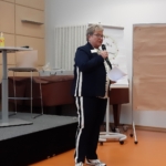 Eine Person in einem marineblauen Anzug mit weißen Streifen spricht in einem hell erleuchteten Raum mit orangefarbenem Bodenbelag in ein Mikrofon und hält Papiere neben einem leeren Flipchart und einem Tisch.