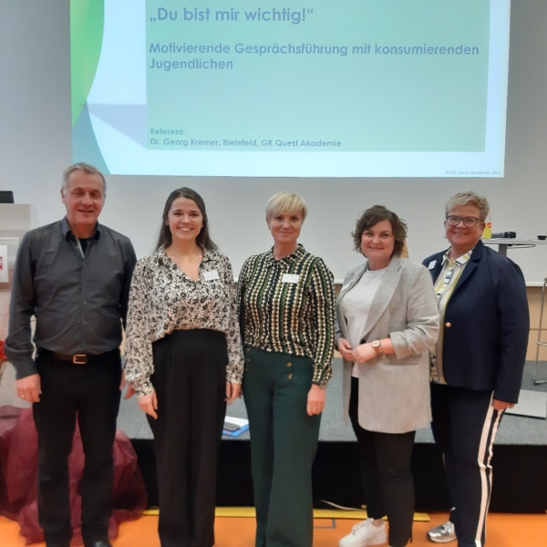 Fünf Personen stehen lächelnd vor einer projizierten Präsentationsfolie in einem Konferenzraum. Die Folie trägt den Titel "Du bist mir wichtig!" und enthält Informationen über Motivationsgespräche mit Jugendlichen.