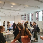 Eine Gruppe von Menschen sitzt an verschiedenen Tischen mit Broschüren und Displays in einem hellen, modernen Raum mit Buntglasfenstern. Einige Personen unterhalten sich, während andere das Material studieren oder eine Erfrischung genießen.