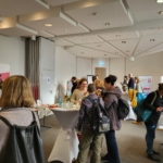 Die Teilnehmer einer Indoor-Networking- oder Informationsveranstaltung mischen sich um Tische mit Broschüren und Displays. An den Wänden sind Plakate und Banner zu sehen, und die Teilnehmer sind in Gespräche vertieft.