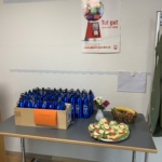 Ein Tisch mit vielen blauen Wasserflaschen, ein Tablett mit belegten Brötchen mit Gurke und Tomate, eine Schale mit Obst, eine kleine Blumenvase und ein Poster an der Wand, auf dem ein Kaugummiautomat abgebildet ist.