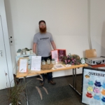 Ein bärtiger Mann mit einem grauen QUERBEET-Hemd steht hinter einem Tisch mit Pflanzen, Blumen, Flaschen und Informationsblättern an einem Veranstaltungsstand. Ein buntes Schild mit QUERBEET hängt am Tisch.