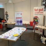 Eine Frau steht bei einer Indoor-Veranstaltung in der Nähe von Tischen, die mit Papieren und Flugblättern bedeckt sind. Zwei Banner sind zu sehen, auf einem steht "Tut gut und klebt nicht". Der Raum hat einen grauen Teppich und eine moderne Deckengestaltung.
