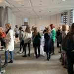 In einem hellen Konferenzraum mit Tischen, Erfrischungsgetränken und Postern tummelt sich eine Gruppe von Menschen und unterhält sich. Einige Teilnehmer stehen in kleinen Gruppen, während andere in der Nähe der Fenster in den Auslagen stöbern.
