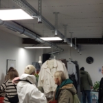 Menschen stöbern in einem modernen, industriell geprägten Geschäft nach Kleidung. Mehrere Regale mit Jacken und Pflanzen sind zu sehen. Die Decke weist freiliegende Rohre und Balken auf, und an der Wand im Hintergrund hängt eine schwarze Wanduhr.