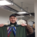 Ein lächelnder Mann mit einer Mütze hält einen bunten, gemusterten Pullover in einem Raum mit Industriebeleuchtung und Kleiderständern im Hintergrund hoch.