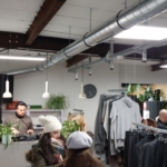 Winterlich gekleidete Kunden stöbern in einem modernen Geschäft mit Pflanzen, Hängelampen und freiliegenden Lüftungsschächten in Regalen mit grauer Kleidung. Ein DJ legt hinter dem Tresen Musik auf.