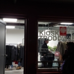 Blick durch ein Fenster in ein Bekleidungsgeschäft mit Kleiderständern und Menschen, die darin stöbern. Ein Schild am Fenster zeigt den Namen "Kiosk Troddwa" und die Öffnungszeiten des Geschäfts an.